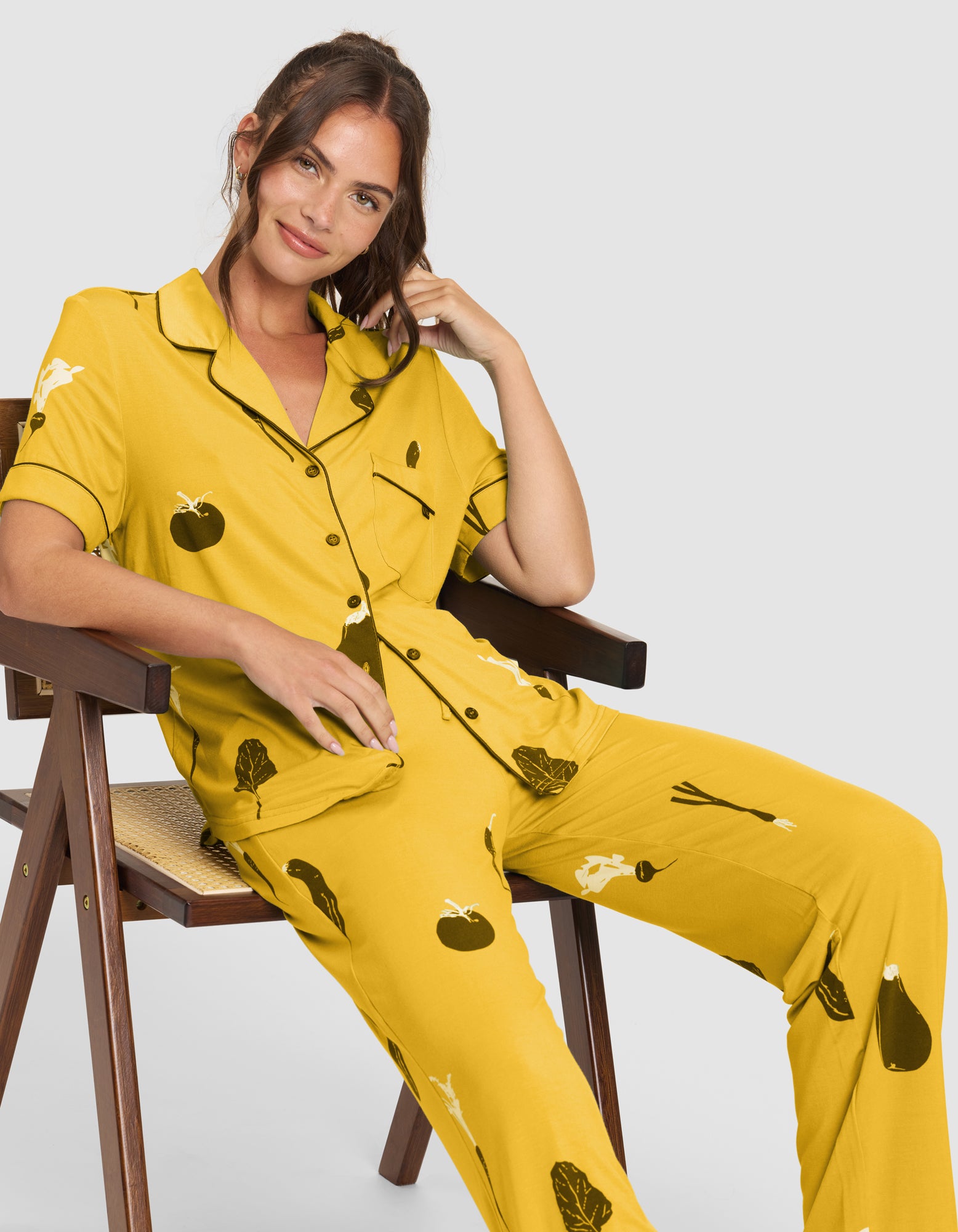 A woman with long brown hair sits on a wooden chair, smiling softly and wearing the HIDE Women's Bamboo Stretch-Knit Pajama Pant (Veggie), a yellow set featuring black and white vegetable illustrations, as she rests her head on her hand. 
