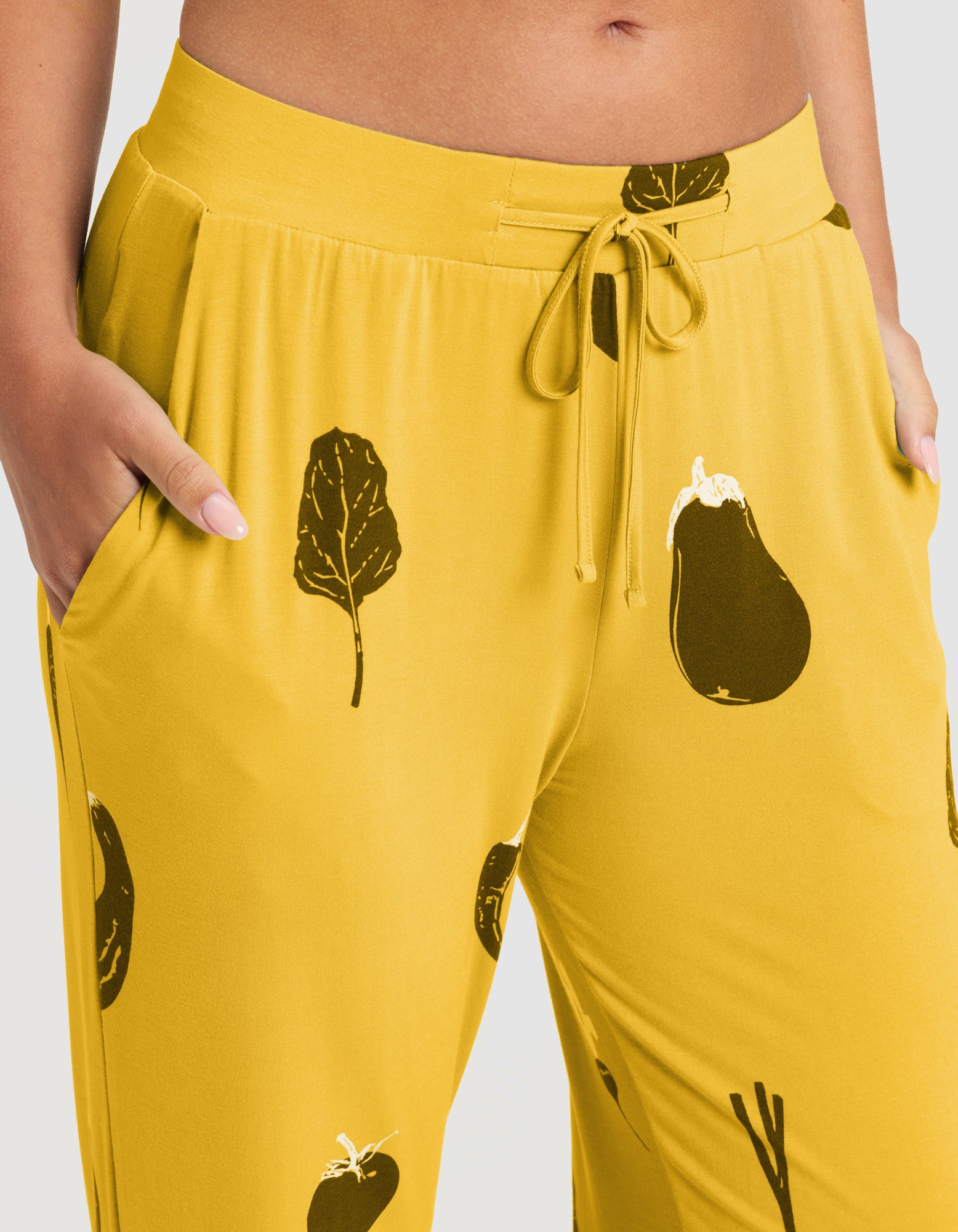 A person wearing HIDE Women's Bamboo Stretch-Knit Pajama Pant (Veggie), featuring bright yellow fabric with black and white vegetable prints, a drawstring waist, and side pockets with hands tucked in. 