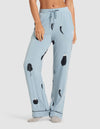 A woman wears HIDE's Women's Bamboo Stretch-Knit Pajama Pant in light blue with black and white vegetable prints, standing barefoot against a plain white background. |Color:Veggies in Smokey Blue
