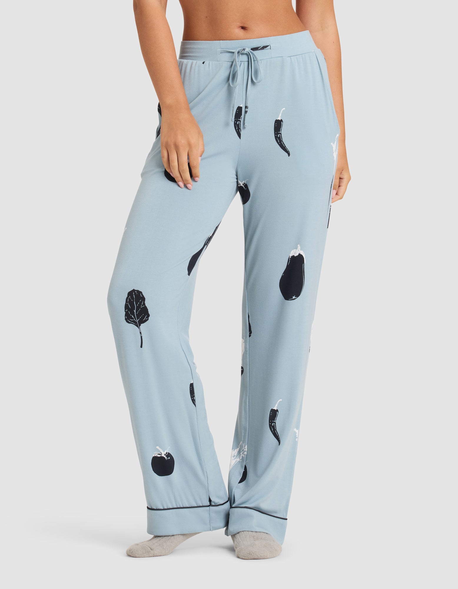 A woman wears HIDE's Women's Bamboo Stretch-Knit Pajama Pant in light blue with black and white vegetable prints, standing barefoot against a plain white background. 