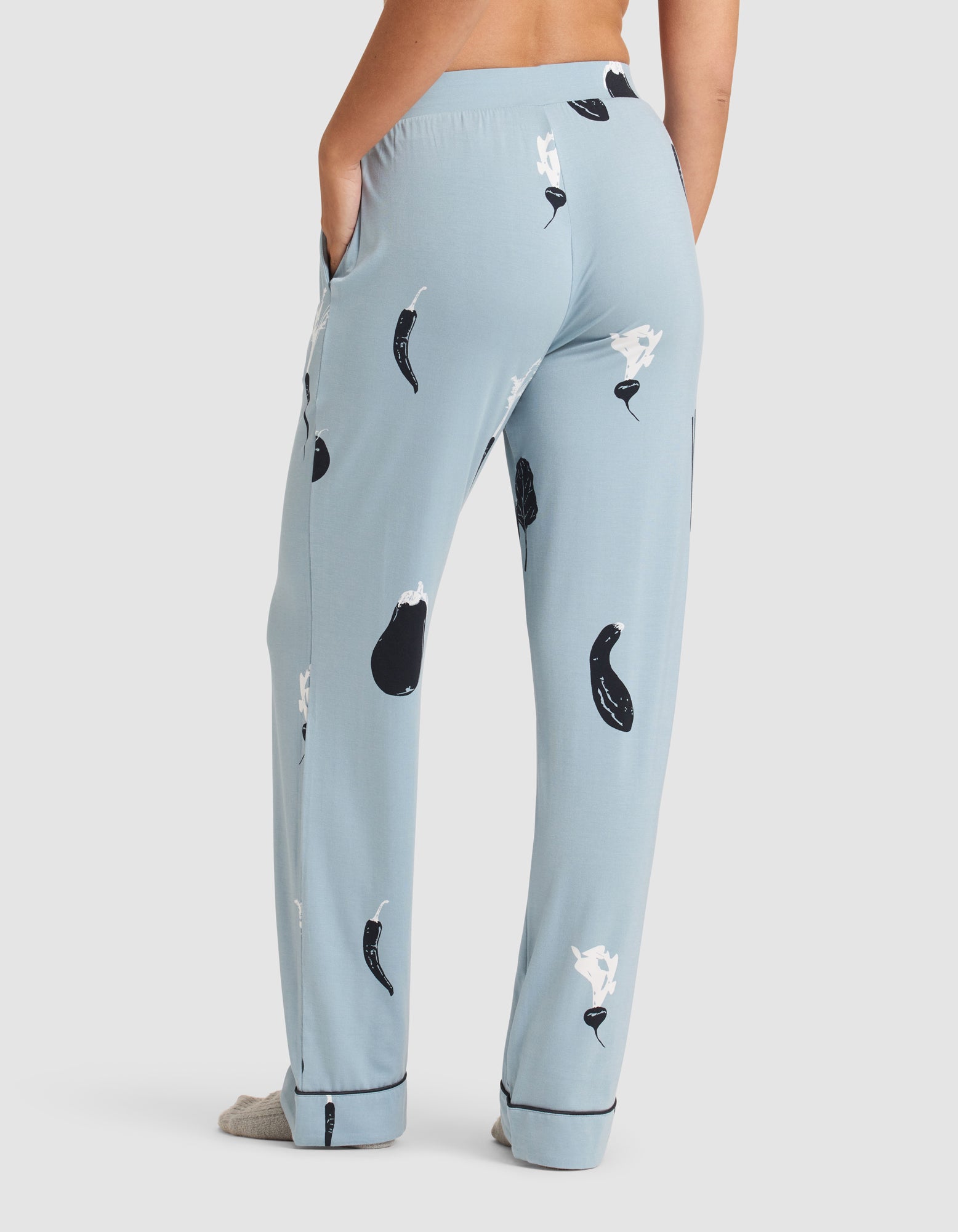 A woman stands with hands in pockets, facing away, wearing HIDE Women's Bamboo Stretch-Knit Pajama Pant in light blue with black and white eggplant and garlic prints, featuring a relaxed fit and cuffed hems. 