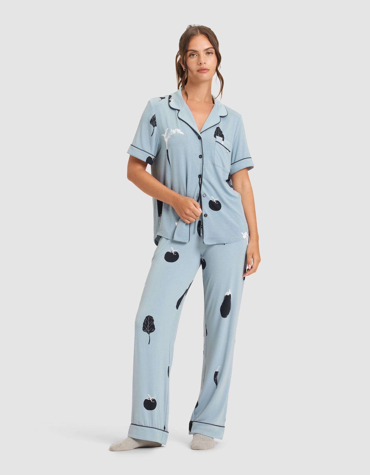 A woman wears HIDE's Women's Bamboo Stretch-Knit Pajama Pant in light blue with black and white fruit and leaf print, paired with gray socks. The set features short sleeves, buttons, a collar, and is shown against a plain white background. 