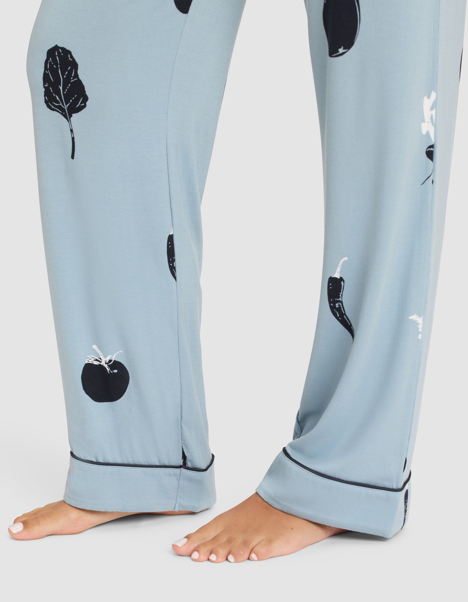 A person wears HIDE Women's Bamboo Stretch-Knit Pajama Pants in light blue with black and white vegetable prints, standing barefoot on a white surface; only the lower legs and feet are visible. 