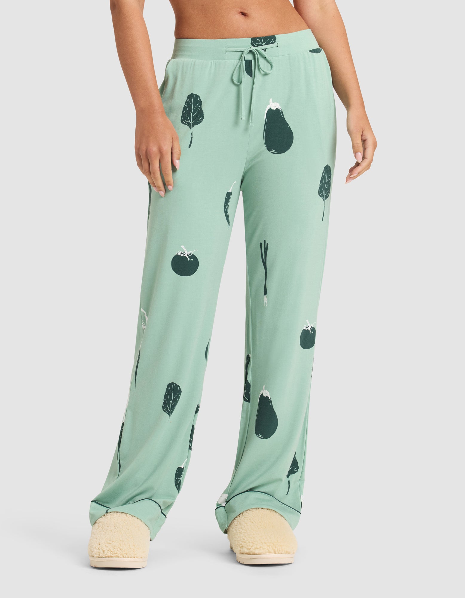 A person wearing HIDE Women's Bamboo Stretch-Knit Pajama Pant in light green with dark green vegetable prints stands with hands by their sides, paired with fuzzy cream slippers. Only the lower body is visible. 