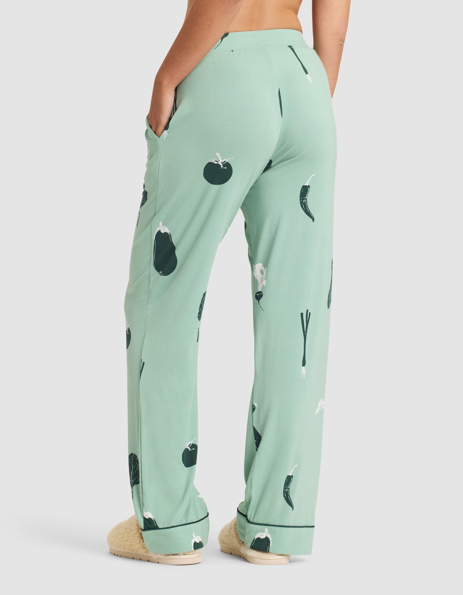 A person wears HIDE's Women's Bamboo Stretch-Knit Pajama Pant in light green with vegetable prints (tomatoes, eggplants, green onions), standing barefoot and viewed from the back with one hand in their pocket. 