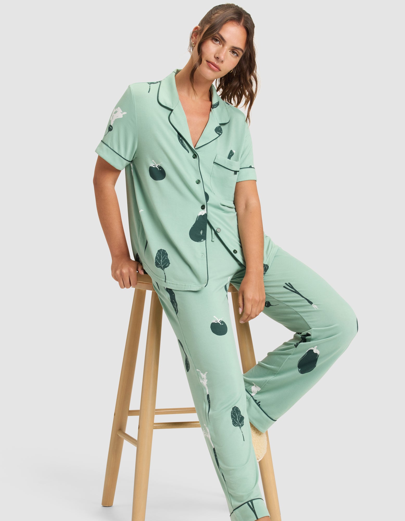 A woman wears the Cozy Earth Women's Bamboo Stretch-Knit Short Sleeve & Pant Pajama Set in mint green with dark green and white vegetable illustrations, sitting sideways on a wooden stool and looking at the camera. 