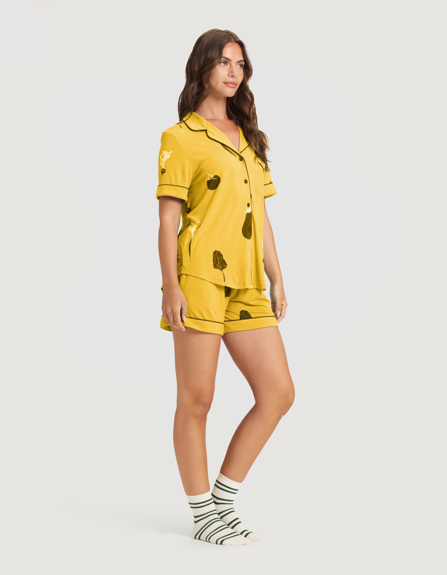 A woman wears the HIDE Women's Bamboo Stretch-Knit Short Sleeve Pajama Top (Veggie) with matching shorts and white socks with green stripes, smiling slightly to the side against a plain light background.