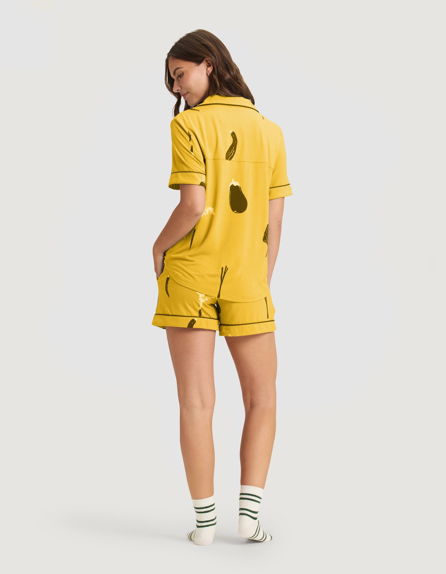 A woman stands with her back to the camera, wearing HIDE's Women's Bamboo Stretch-Knit Short Sleeve Pajama Top (Veggie) and white ankle socks with green stripes.