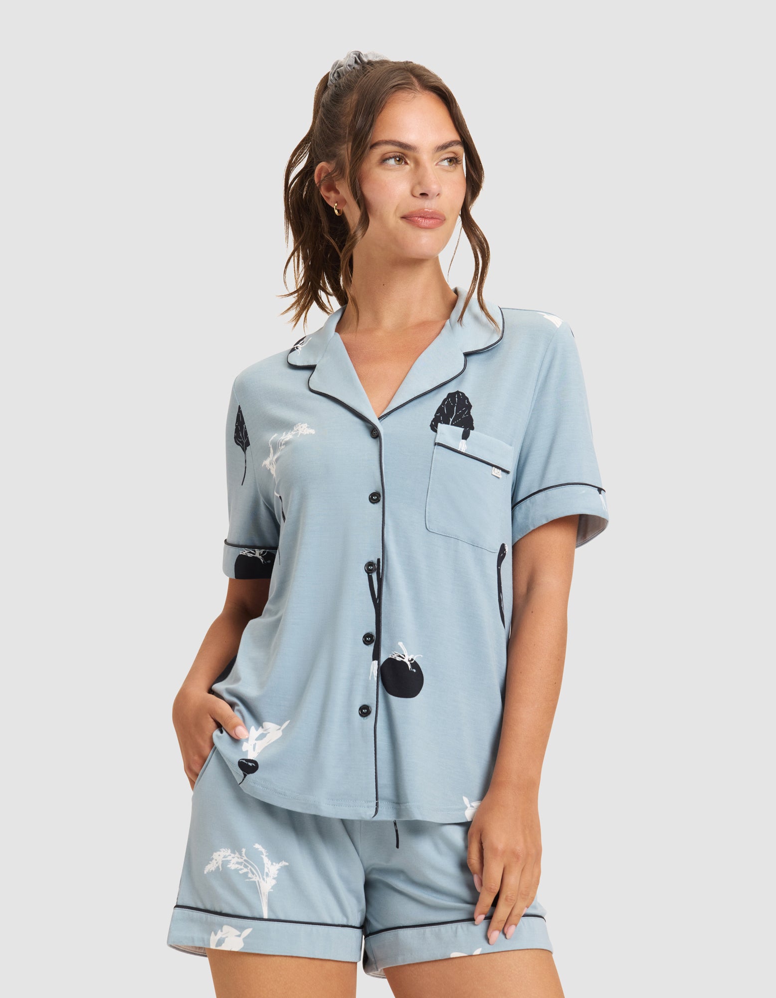 A woman wears the HIDE Women's Bamboo Stretch-Knit Short Sleeve Pajama Top in light blue with a white and black abstract pattern, paired with matching shorts. She stands with one hand in her pocket, looking to the side. |Color:Veggies in Smokey Blue