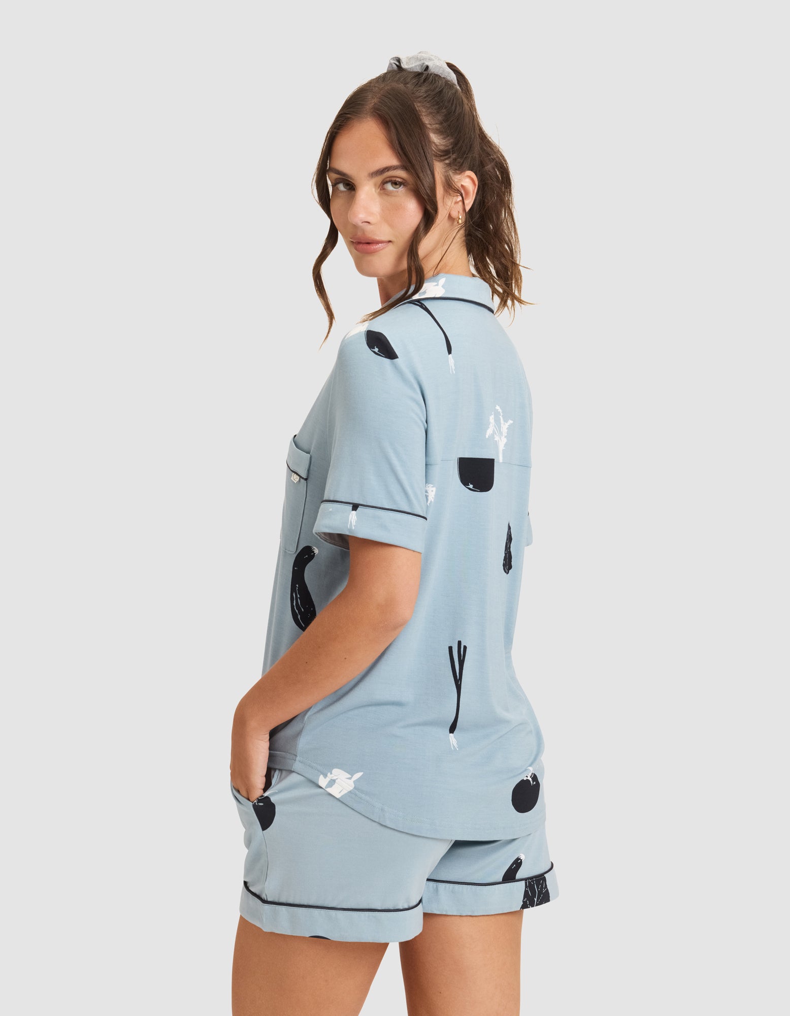 A woman with wavy brown hair in a ponytail models HIDE's Women's Bamboo Stretch-Knit Short Sleeve Pajama Top in light blue with black and white abstract patterns, posing with one hand in her pocket and looking over her shoulder. 