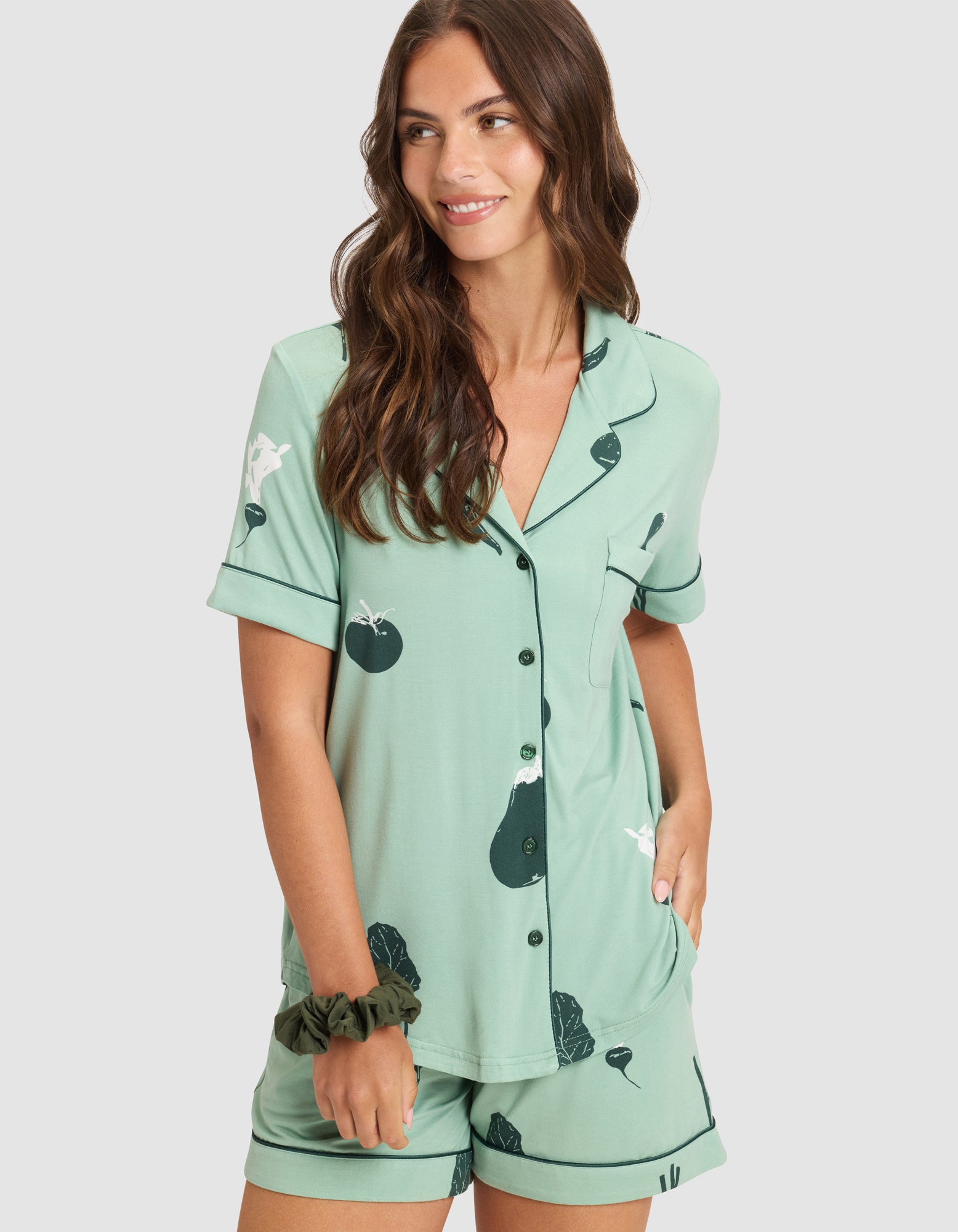 A woman with long brown hair wears the HIDE Women's Bamboo Stretch-Knit Short Sleeve Pajama Top and matching shorts with a leafy vegetable print. She smiles, one hand in her pocket, against a plain light background. |Color:Veggies in Wintergreen