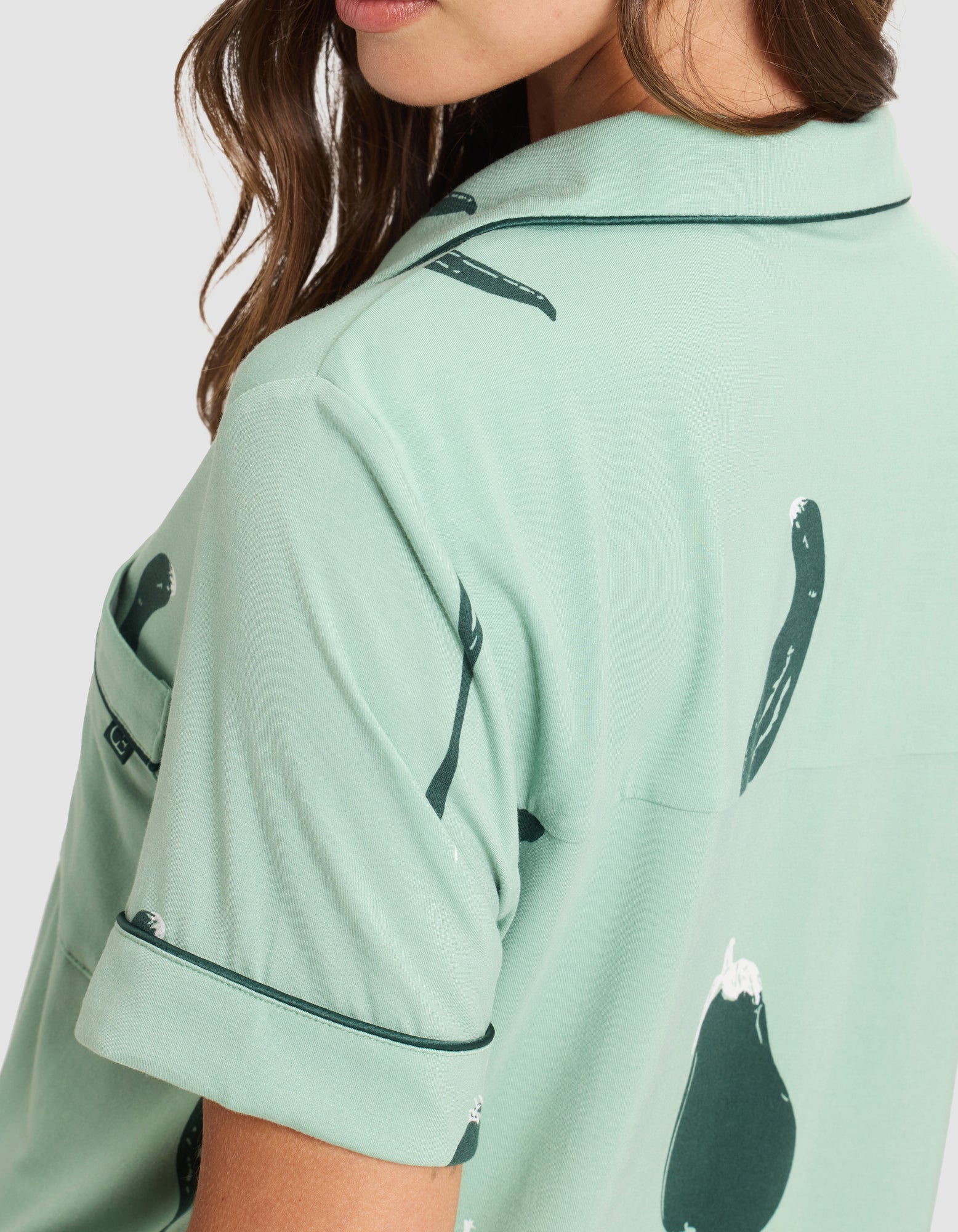 A woman wears the HIDE Women's Bamboo Stretch-Knit Short Sleeve Pajama Top, light green with dark green eggplant and pea pod print, collar, piped short sleeves, and chest pocket. Only her lower face and upper body are visible. 