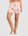 A person stands barefoot in HIDE Women's Bamboo Stretch-Knit Pajama Shorts, featuring a light pink color with a drawstring and brown leaf and vegetable prints, against a plain light background. |Color:Veggies in Dusty Rose