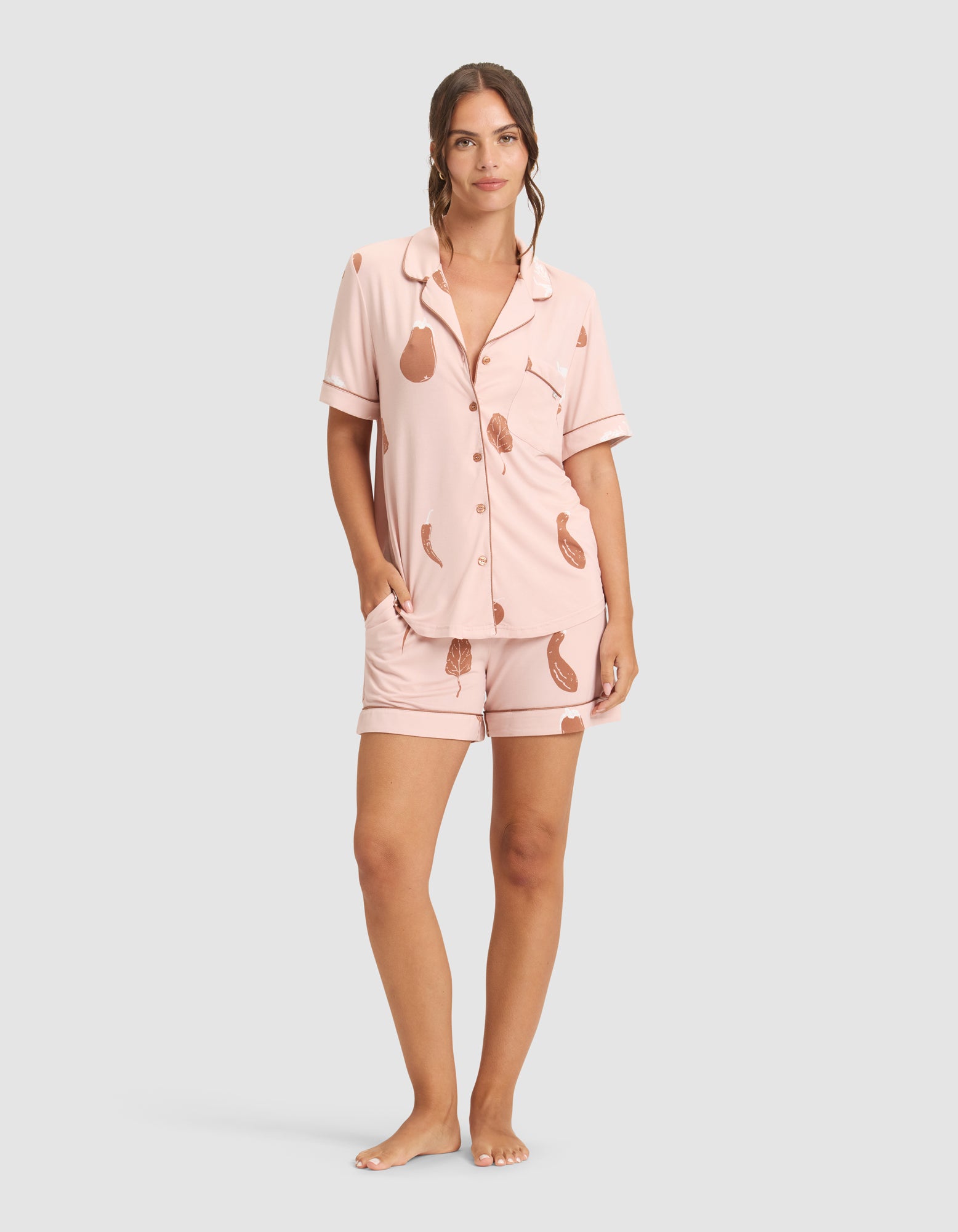 A woman stands barefoot against a plain background, smiling slightly and wearing HIDE Women's Bamboo Stretch-Knit Pajama Shorts with a light pink, short-sleeved, button-up top and brown pear pattern. Her left hand is in her pocket. 