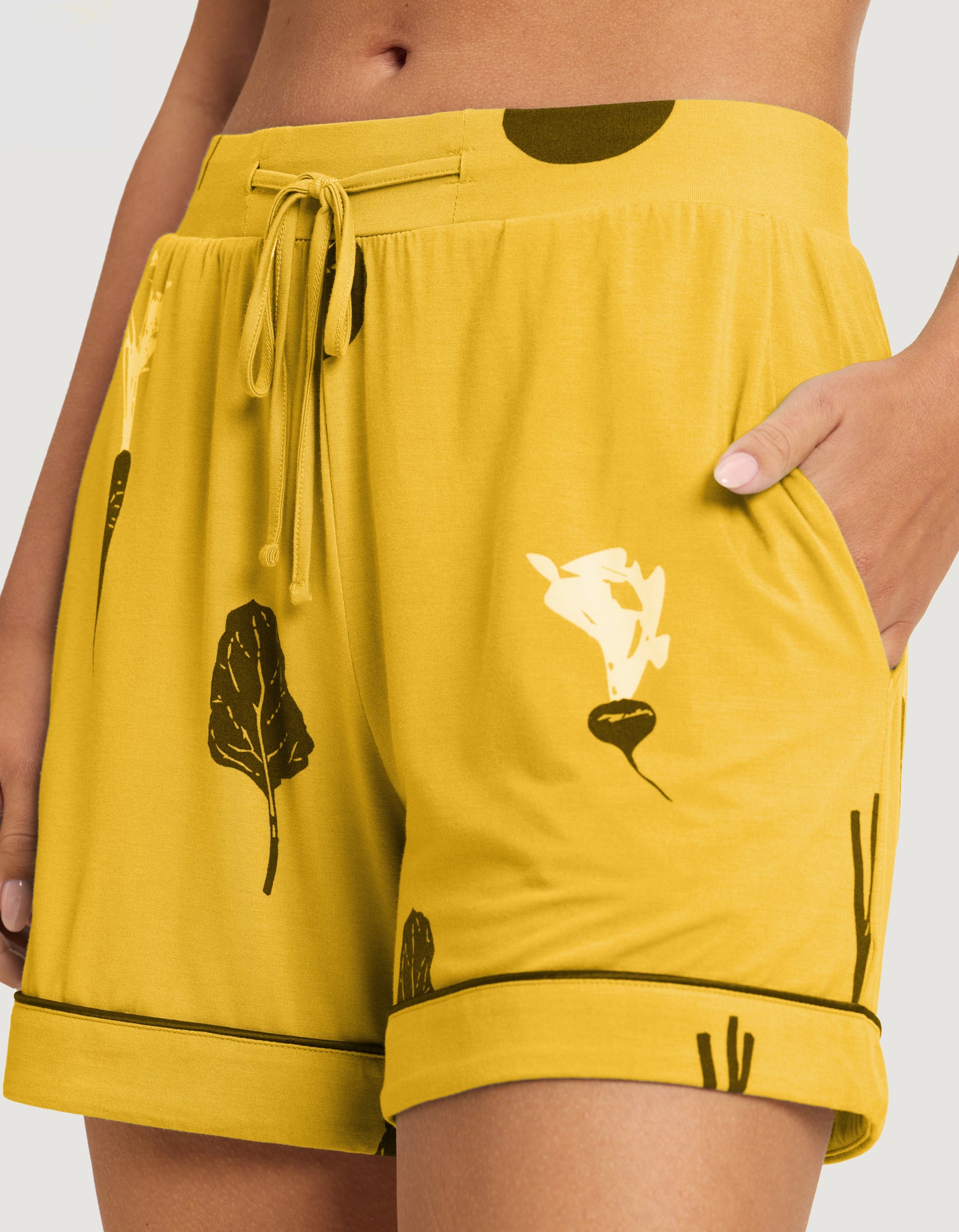 A person stands with one hand in their pocket, wearing HIDE’s Last Chance Women's Bamboo Stretch-Knit Pajama Shorts (Veggie) featuring a yellow base with black and white vegetable prints, drawstring waist, and rolled cuffs. 