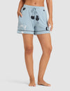 A person stands barefoot against a plain white background, wearing HIDE Women's Bamboo Stretch-Knit Pajama Shorts in light blue with a black and white botanical print, drawstring waist, and cuffed hems. |Color:Veggies in Smokey Blue