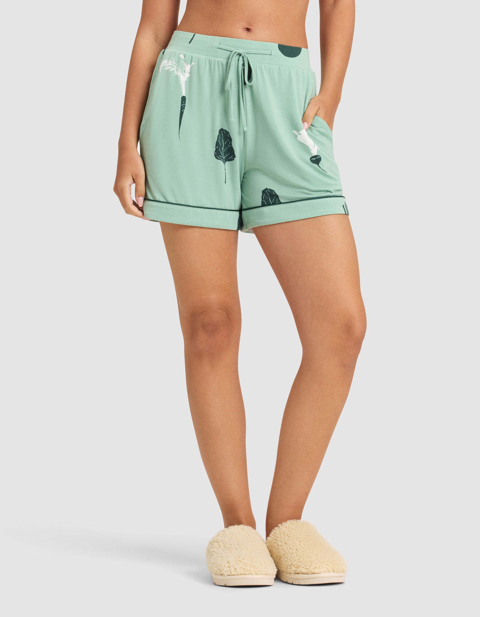 A person wears HIDE Women's Bamboo Stretch-Knit Pajama Shorts in light green with a leaf print, drawstring waist, and black trim, paired with cream fuzzy slippers. Upper body not visible. 