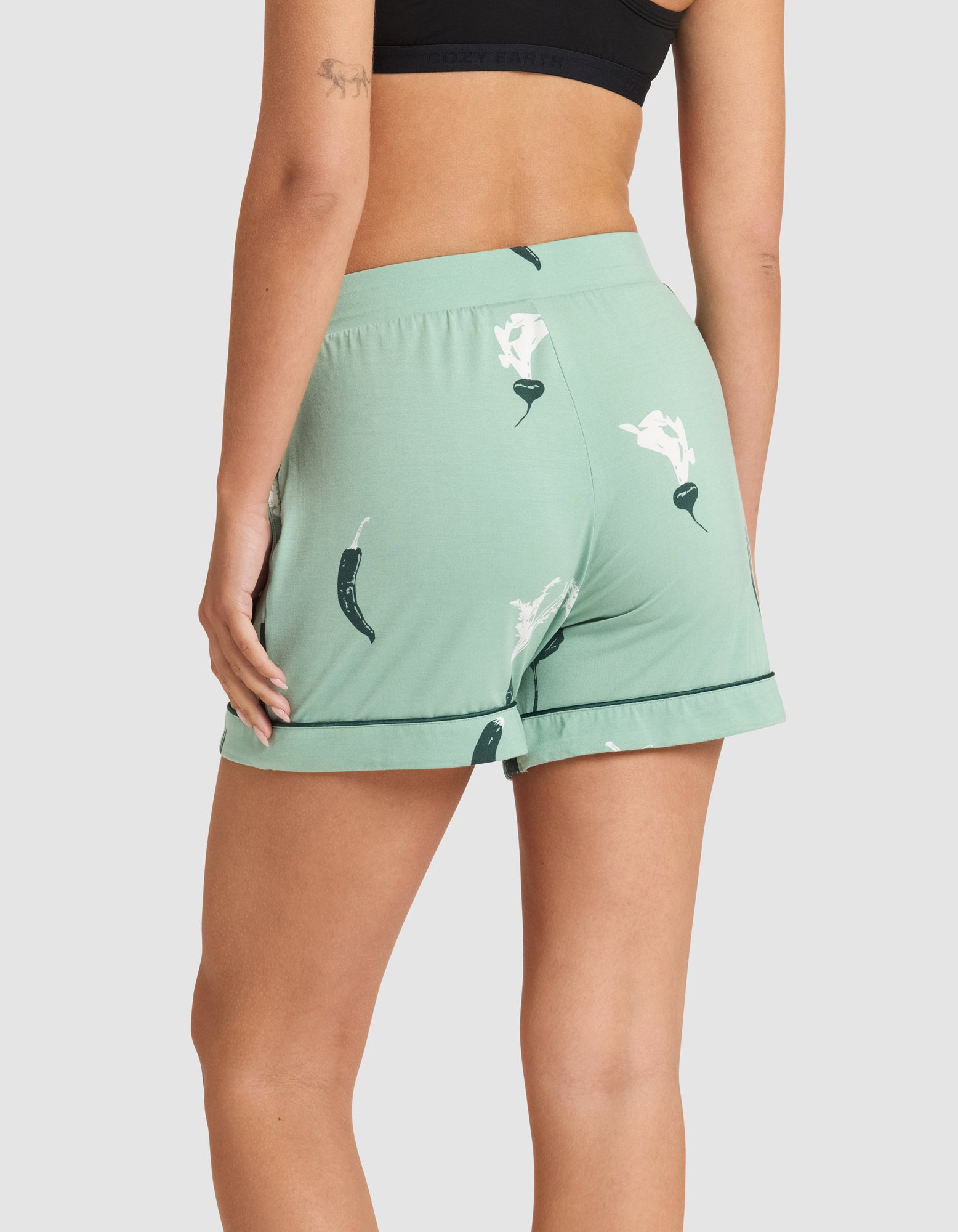 A person is shown from the back wearing HIDE Women's Bamboo Stretch-Knit Pajama Shorts in light green with a chili pepper print and a cuffed hem, paired with a black sports bra. The shorts have a loose fit. A small arm tattoo is visible. 