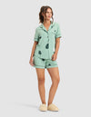 A woman in a relaxed pose wears HIDE's Women's Bamboo Stretch-Knit Pajama Short set in mint green with a leaf print, paired with beige fuzzy slippers. Her brown hair is styled in a high ponytail. |Color:Veggies in Wintergreen