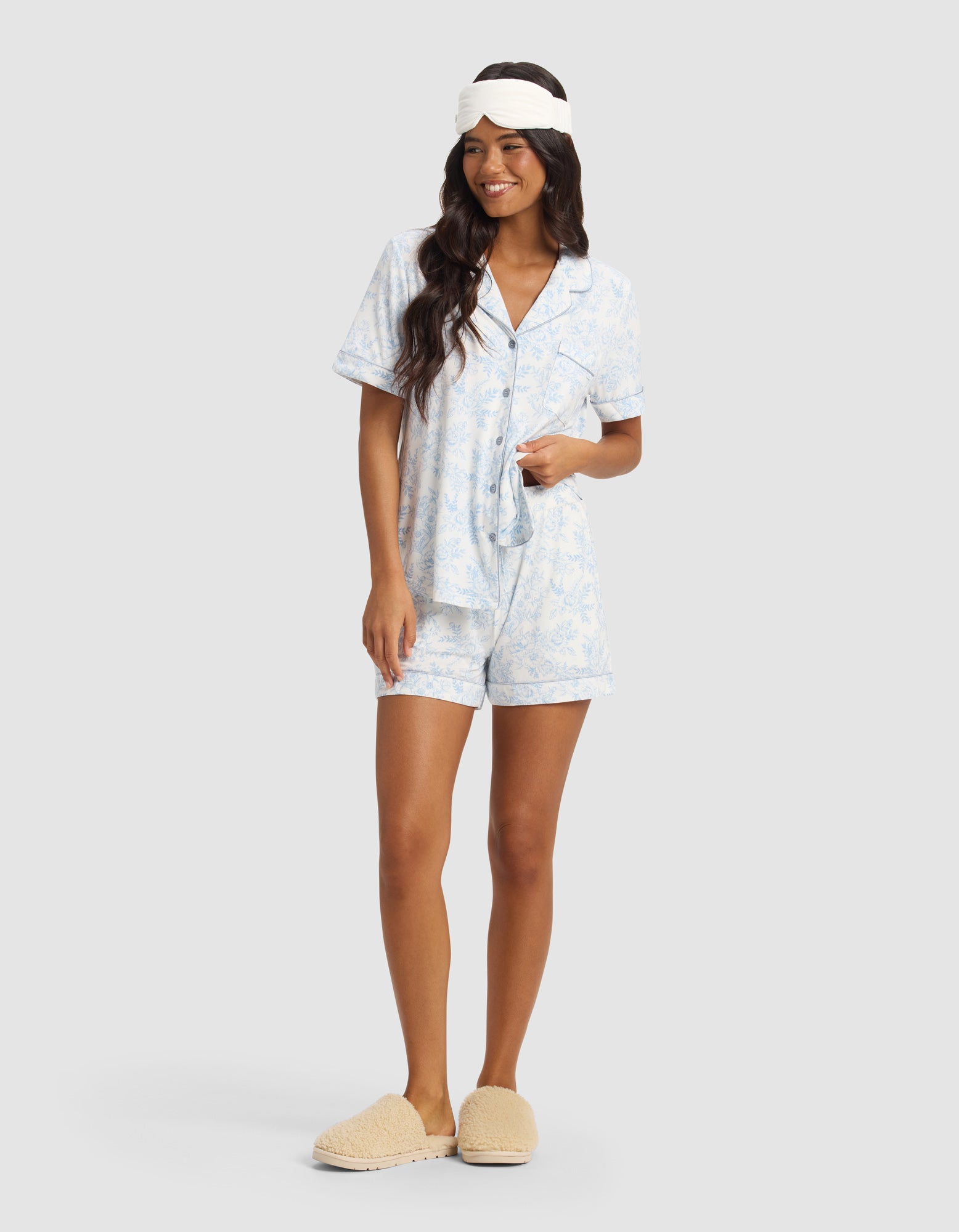 A woman smiles against a plain light background, wearing Cozy Earth's Women's Bamboo Stretch-Knit Pajama Short set in light blue with a white sleep mask and beige slippers.
