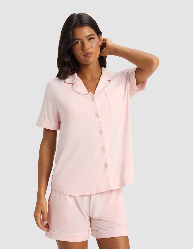 A woman wears the Cozy Earth Women's Bamboo Stretch-Knit Short Sleeve Pajama Top in light pink with matching shorts, standing against a plain light gray background, one hand touching her hair.