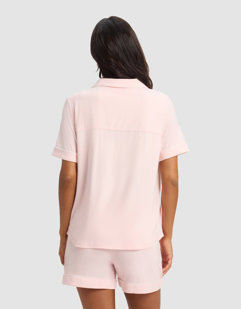 A woman with long dark hair faces away, wearing the Cozy Earth Women's Bamboo Stretch-Knit Short Sleeve Pajama Top in light pink and matching shorts, set against a plain light gray background.