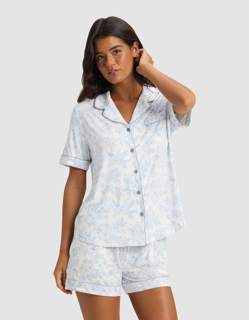 A woman with long dark hair models the Cozy Earth Women's Bamboo Stretch-Knit Short Sleeve Pajama Top in light blue and white with a floral pattern and buttons, standing against a plain light gray background.