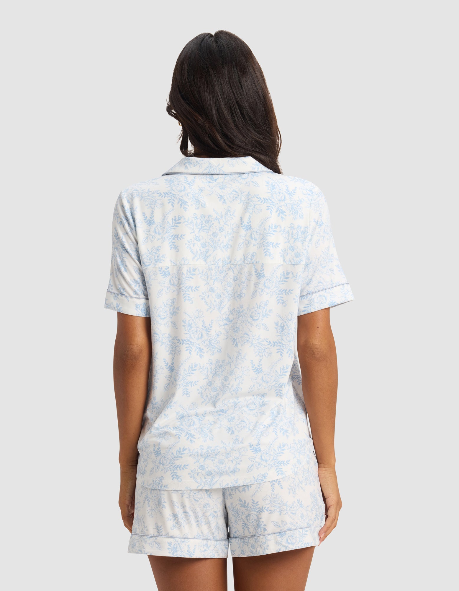 A woman with long dark hair stands facing away, wearing the Cozy Earth Women's Bamboo Stretch-Knit Short Sleeve Pajama Top in light blue and white floral, paired with shorts, against a plain gray background.