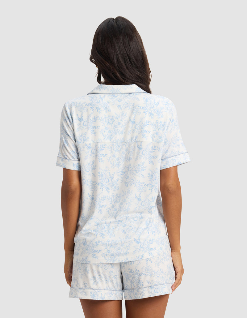 A woman with long dark hair stands facing away, wearing the Cozy Earth Women's Bamboo Stretch-Knit Short Sleeve Pajama Top in light blue and white floral, paired with shorts, against a plain gray background.