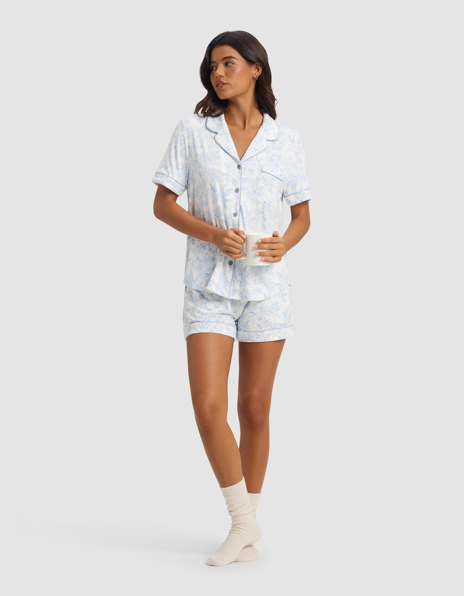 A woman wears the Cozy Earth Women's Bamboo Stretch-Knit Short Sleeve Pajama Top in light blue and white, paired with matching shorts and cream-colored socks, holding a white mug and looking to the side.