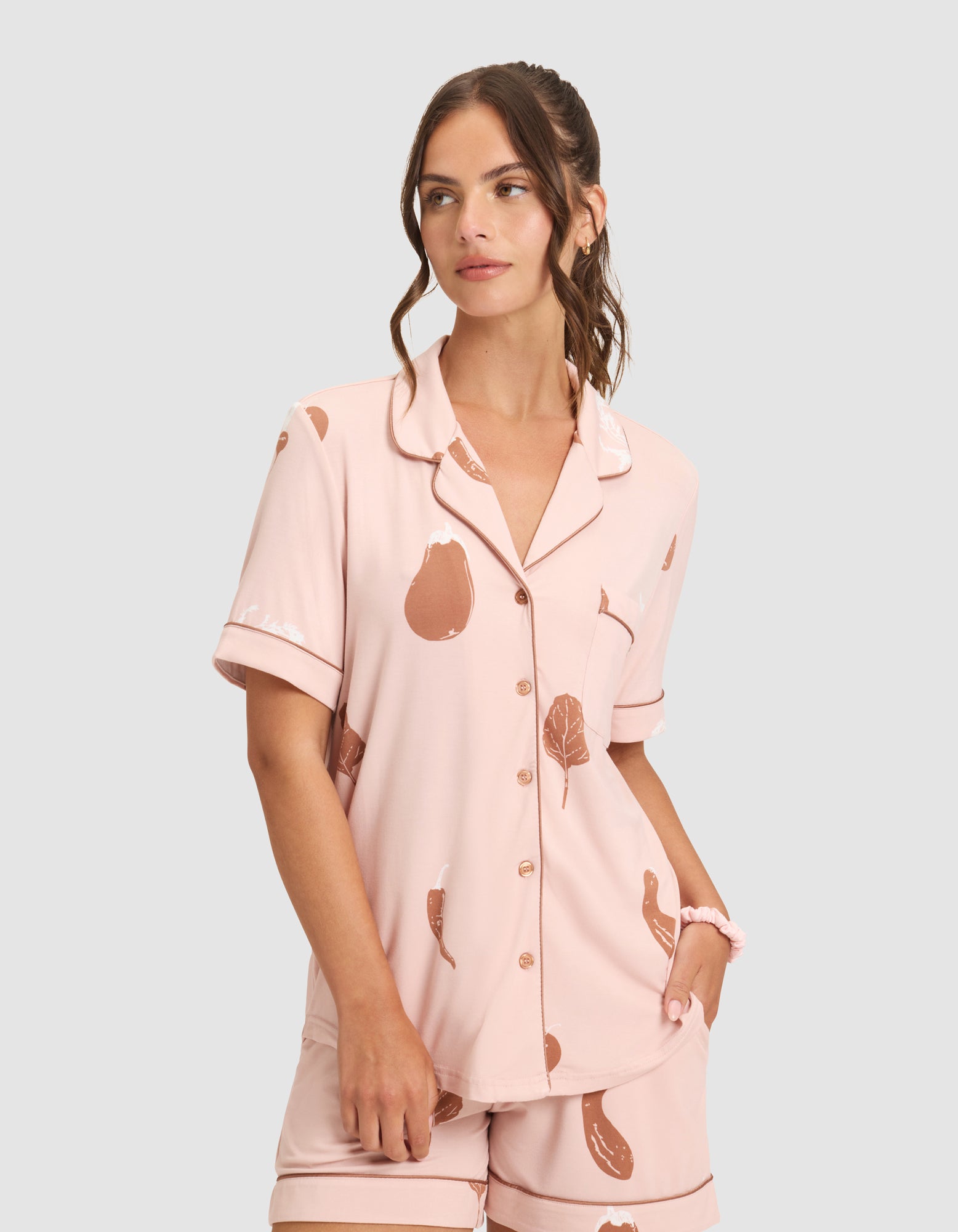 A woman wears the HIDE Women's Bamboo Stretch-Knit Short Sleeve Pajama Top in light pink with brown vegetable prints, standing with one hand in her pocket against a plain gray background. 