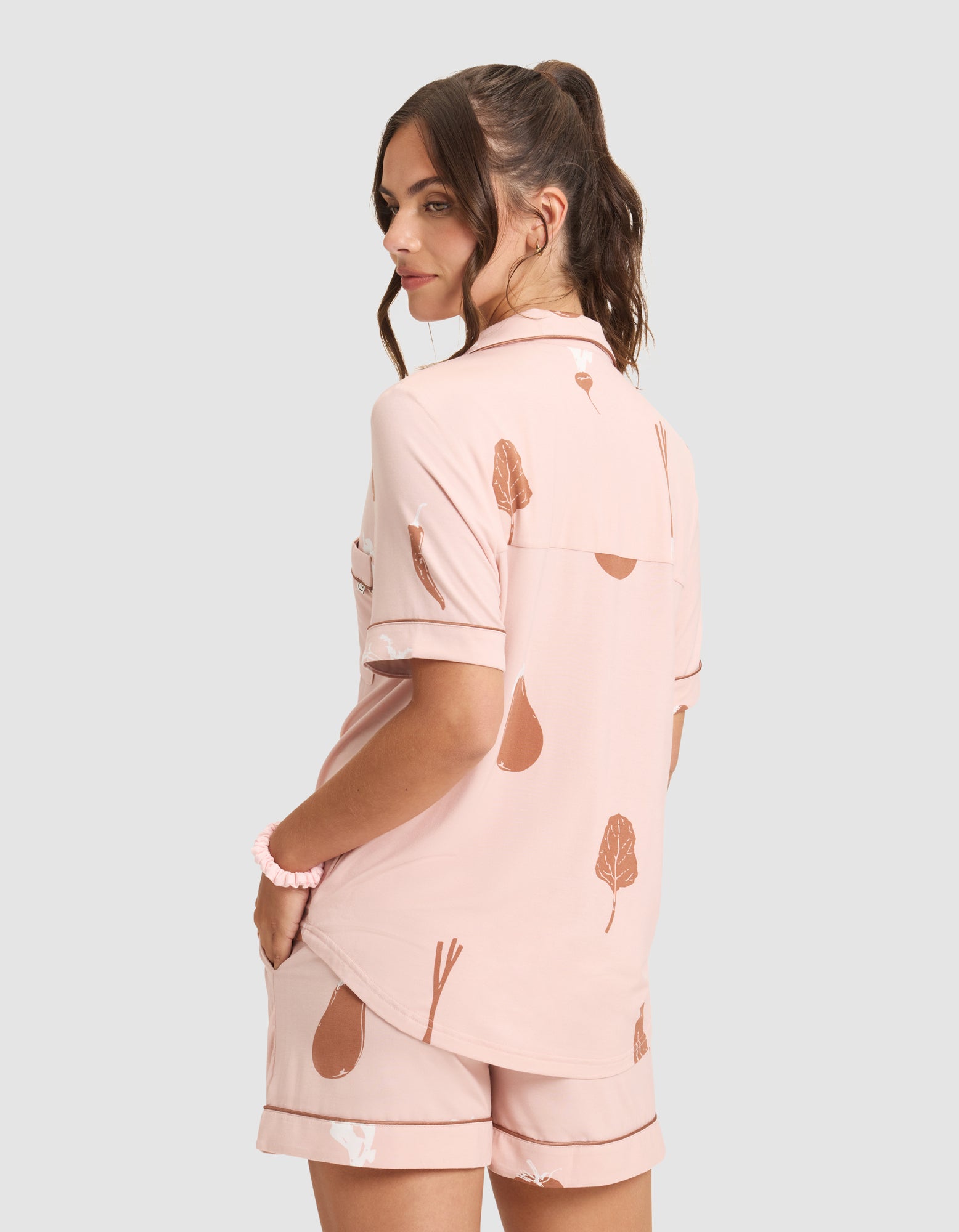 A woman with brown hair in a ponytail wears the HIDE Women's Bamboo Stretch-Knit Short Sleeve Pajama Top, light pink with brown fruit and vegetable prints, seen from the back with her hands in her shorts pockets, looking over her shoulder. 