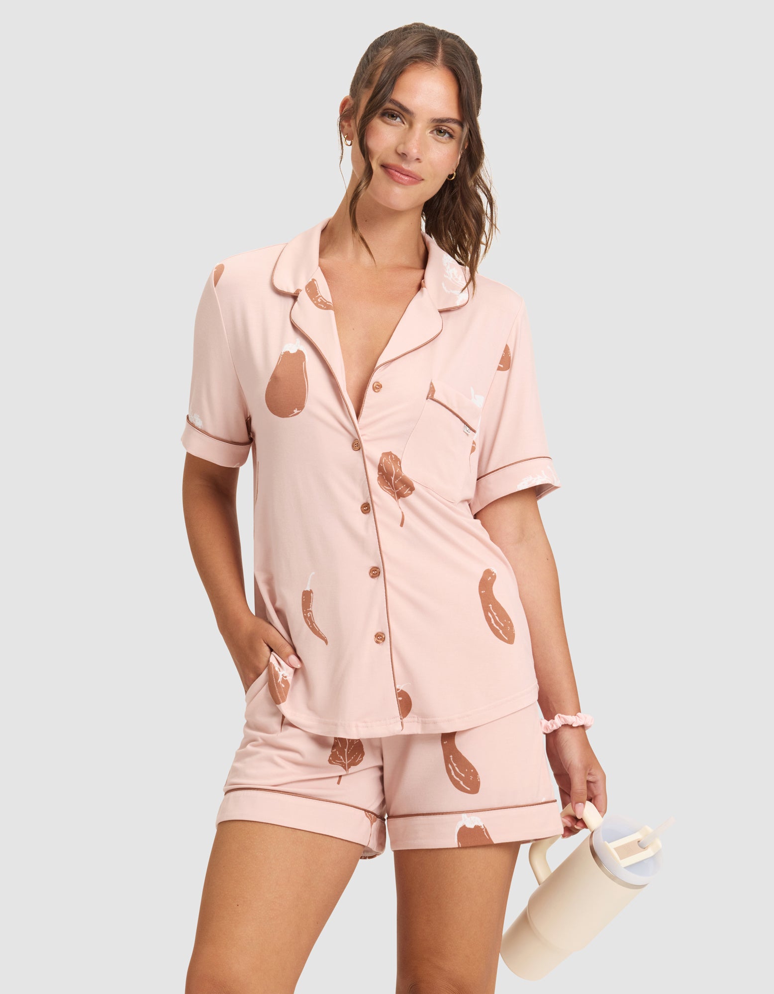 A woman in HIDE's Women's Bamboo Stretch-Knit Short Sleeve Pajama Top, light pink with an eggplant print, stands smiling against a plain background, one hand in her pocket and holding a cup with the other. 