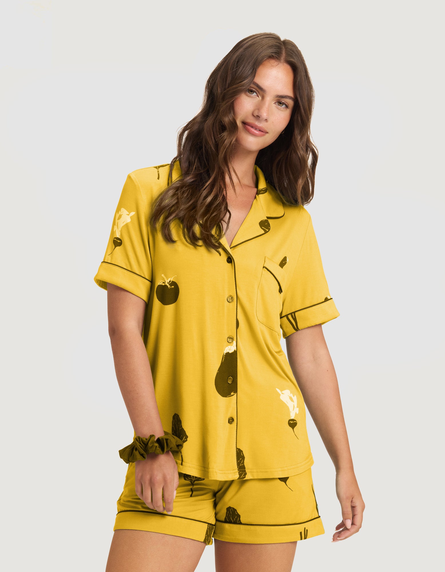 A woman with long brown hair poses against a light background in Women’s Bamboo Stretch-Knit Short Sleeve Pajama Top (Veggie) and matching shorts, both yellow with black abstract prints. She wears a matching scrunchie on her wrist. |Color:Veggies in Mango