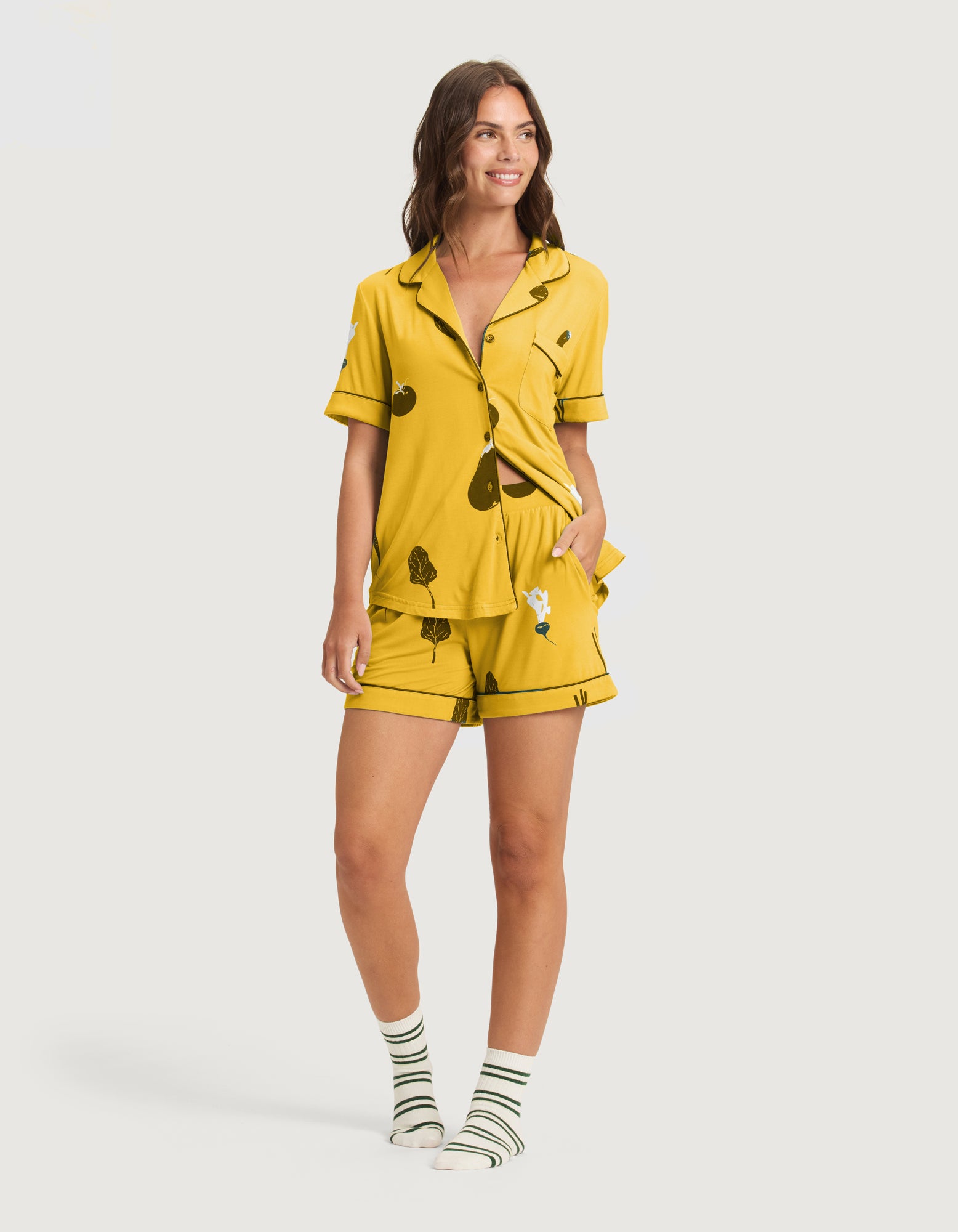 A woman smiles in Cozy Earth's Women's Bamboo Stretch-Knit Short Sleeve Pajama Set (Veggie), featuring a fun veggie print, matching shorts, and striped socks. Her hand is in her pocket as she stands against a plain white background. |Color:Veggies in Mango