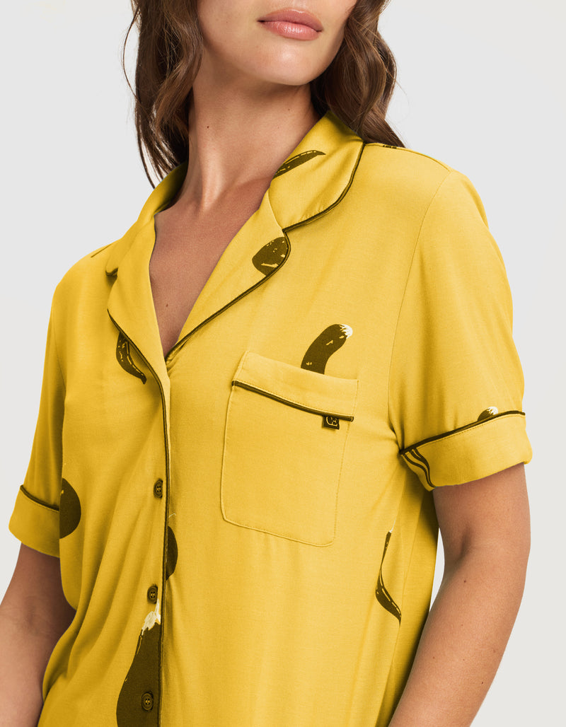A woman wears Cozy Earth's Women's Bamboo Stretch-Knit Short Sleeve Pajama Set (Veggie), featuring a mustard yellow button-up top with black piping, abstract black pattern, chest pocket, and a relaxed fit. Her face is partially visible.