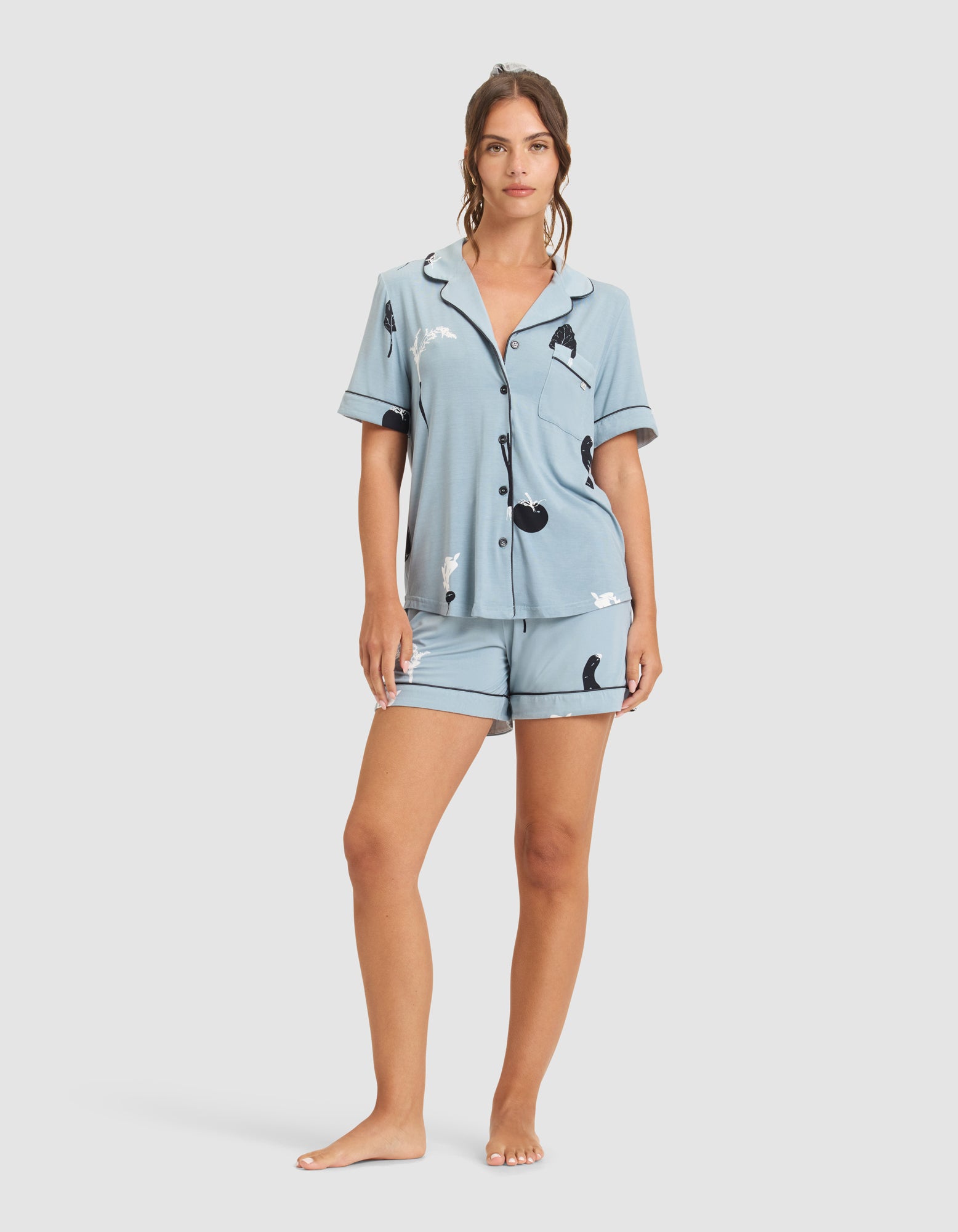 A woman stands barefoot facing forward against a white background, wearing the HIDE Women's Bamboo Stretch-Knit Short Sleeve Pajama Top and matching shorts, both featuring a playful black and white animal print. |Color:Veggies in Smokey Blue