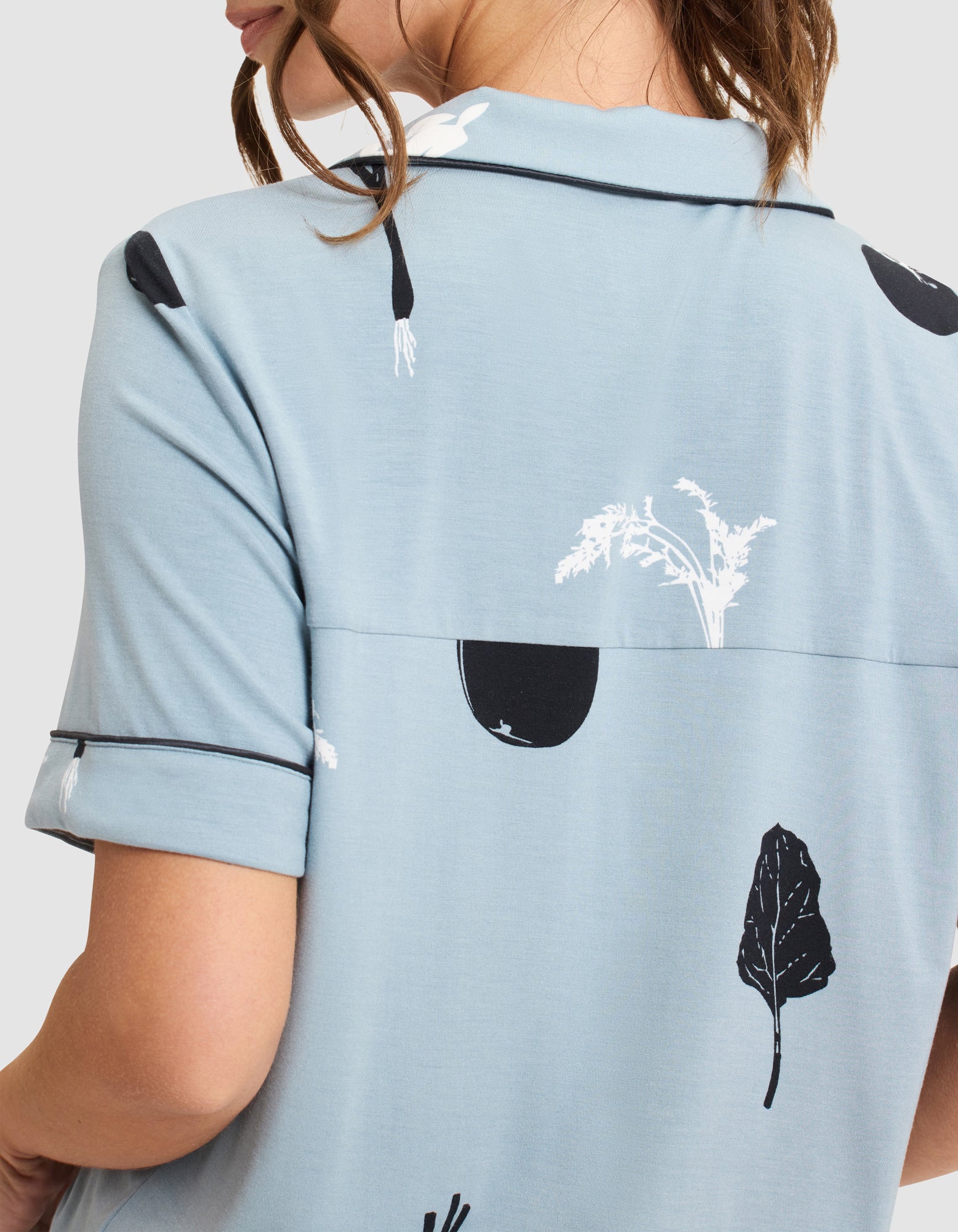 A person is seen from the back wearing HIDE's Women's Bamboo Stretch-Knit Short Sleeve Pajama Top in light blue with a collar, black piping, and a white and black vegetable print. 