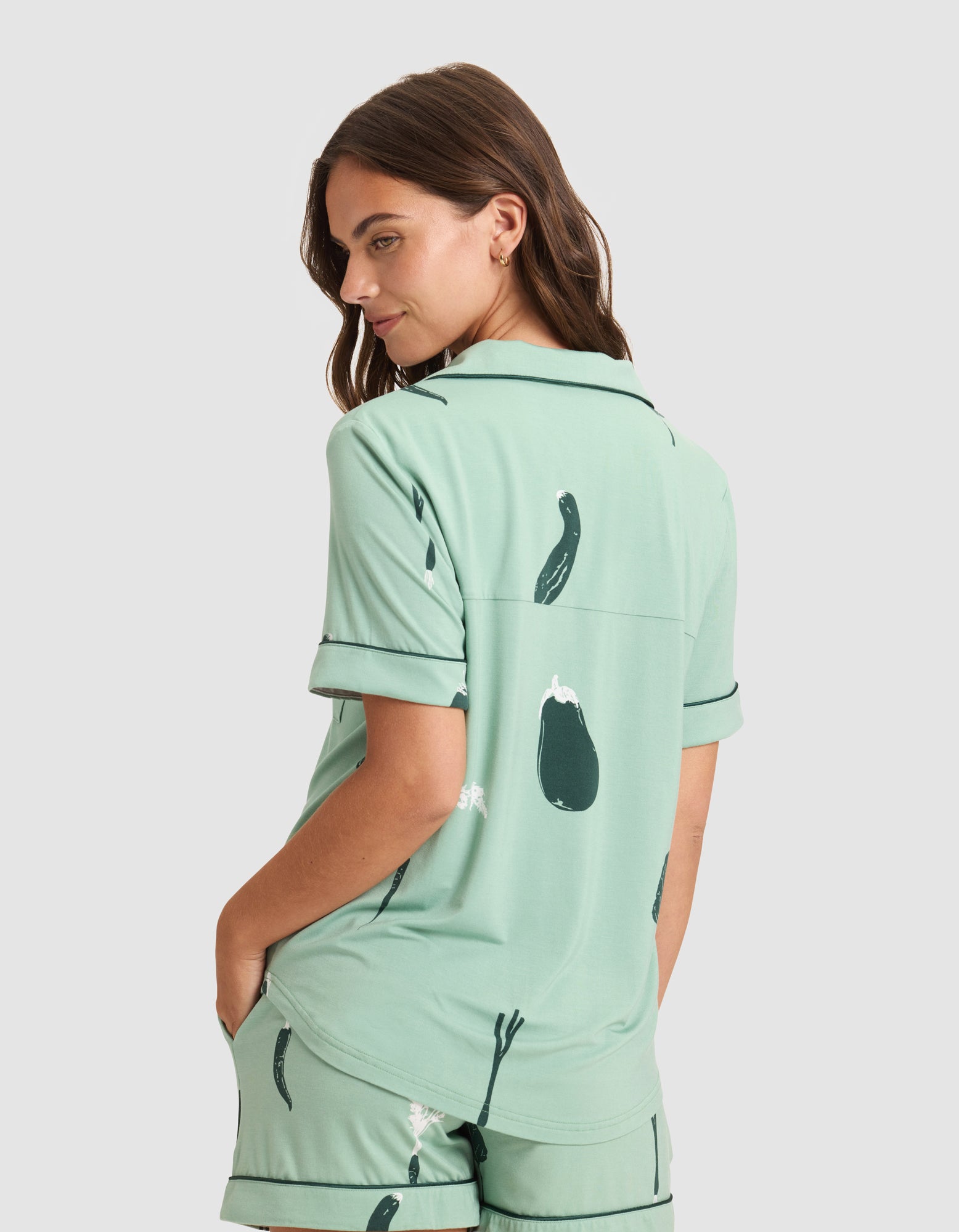 A woman faces away from the camera in a HIDE Women's Bamboo Stretch-Knit Short Sleeve Pajama Top, light green with dark green eggplant print. She has her hands in her shorts pockets and looks over her shoulder against a neutral background. 