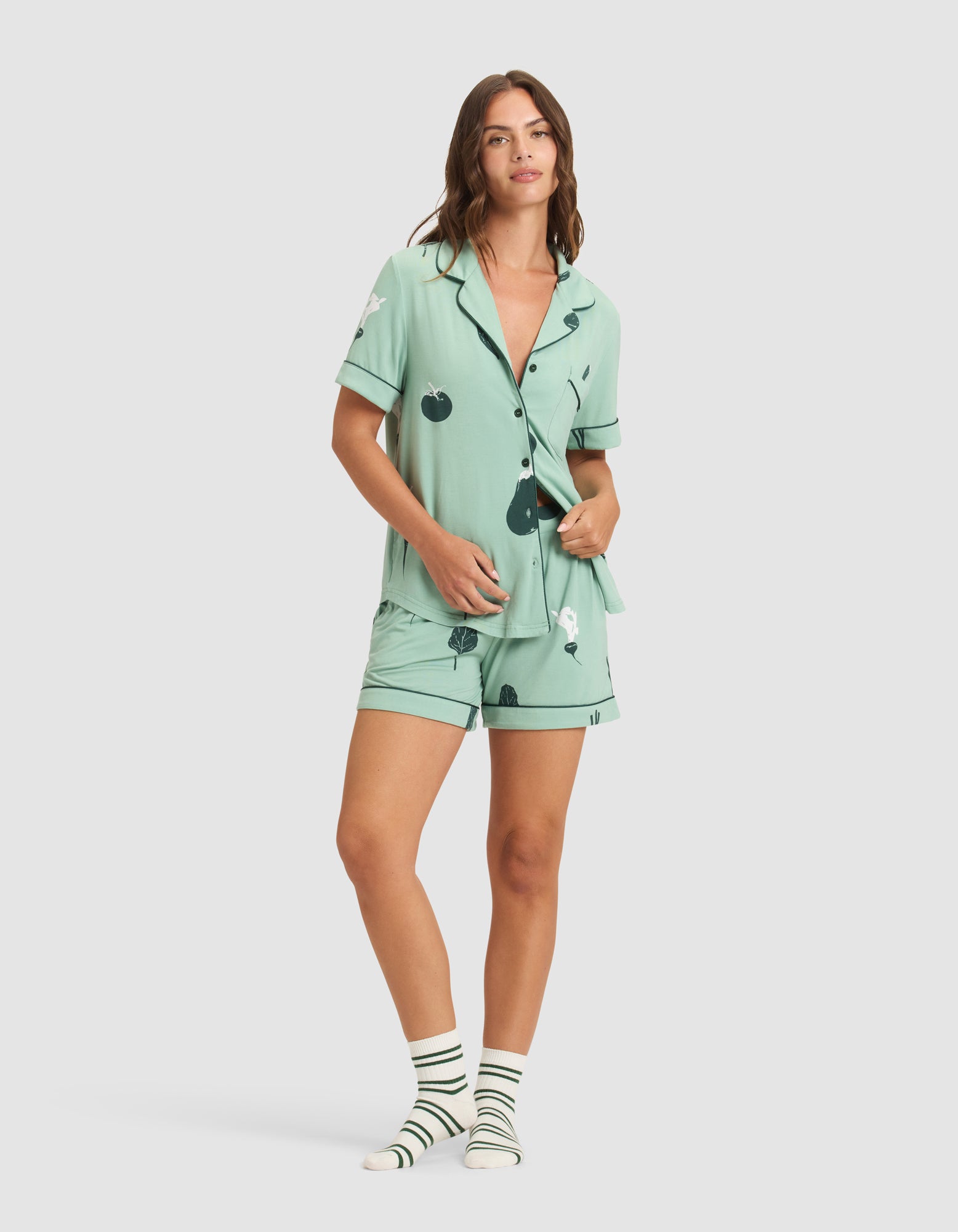 A woman wears HIDE's Women's Bamboo Stretch-Knit Short Sleeve Pajama Top in light green with a cherry print, paired with matching shorts and striped white and green socks, standing against a plain light background. 