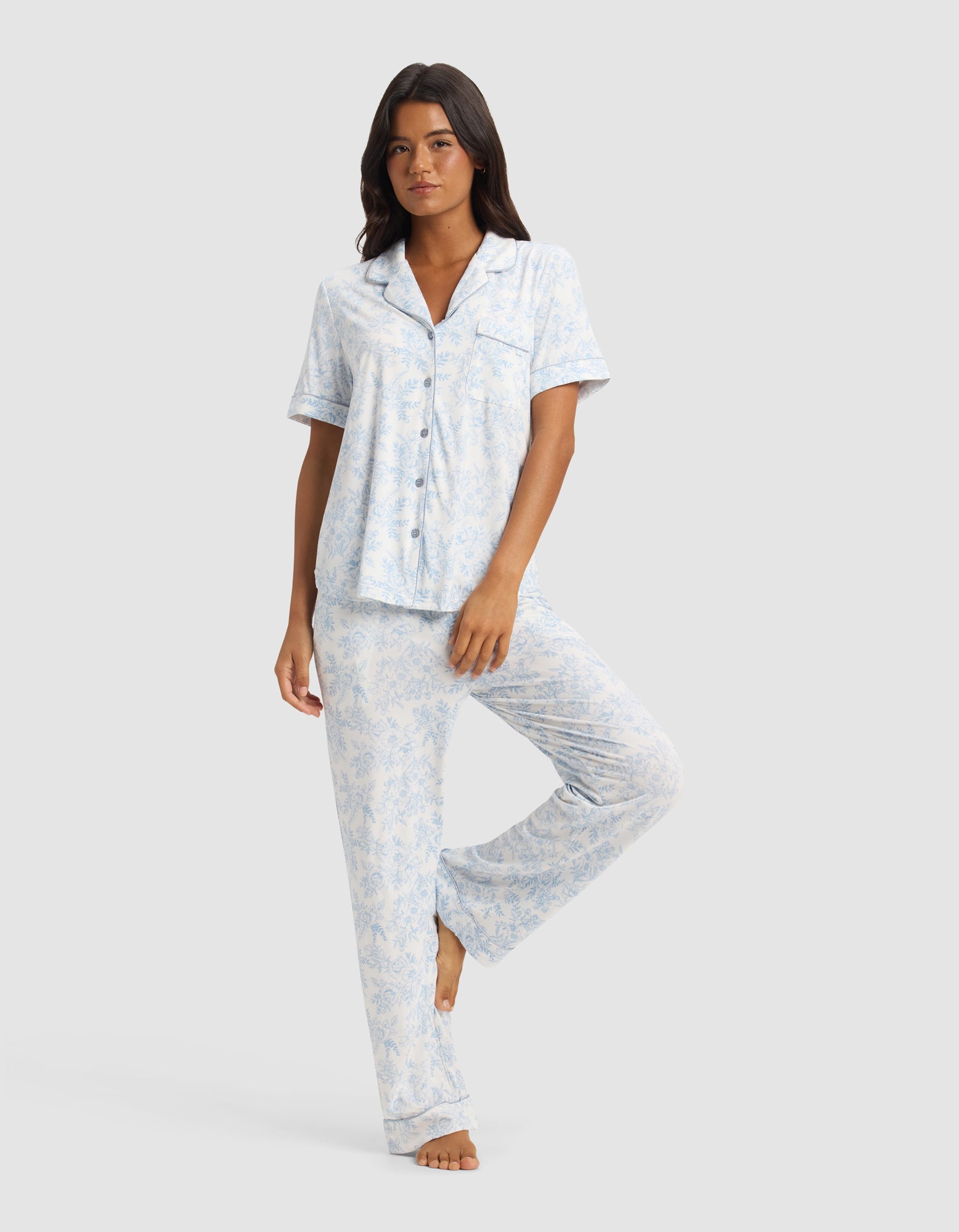 A woman poses barefoot against a plain light background, wearing the Cozy Earth Women's Bamboo Stretch-Knit Short Sleeve & Pant Pajama Set in a light blue and white pattern.