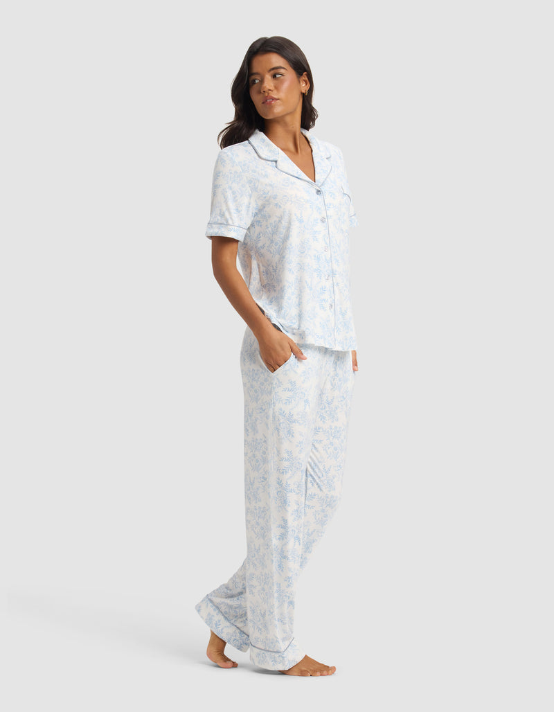 A woman stands barefoot against a plain background, wearing the Cozy Earth Women's Bamboo Stretch-Knit Short Sleeve & Pant Pajama Set in light blue and white. The set features a collared, button-up top with chest pocket and matching pants. She looks to the side with hands in her pockets.