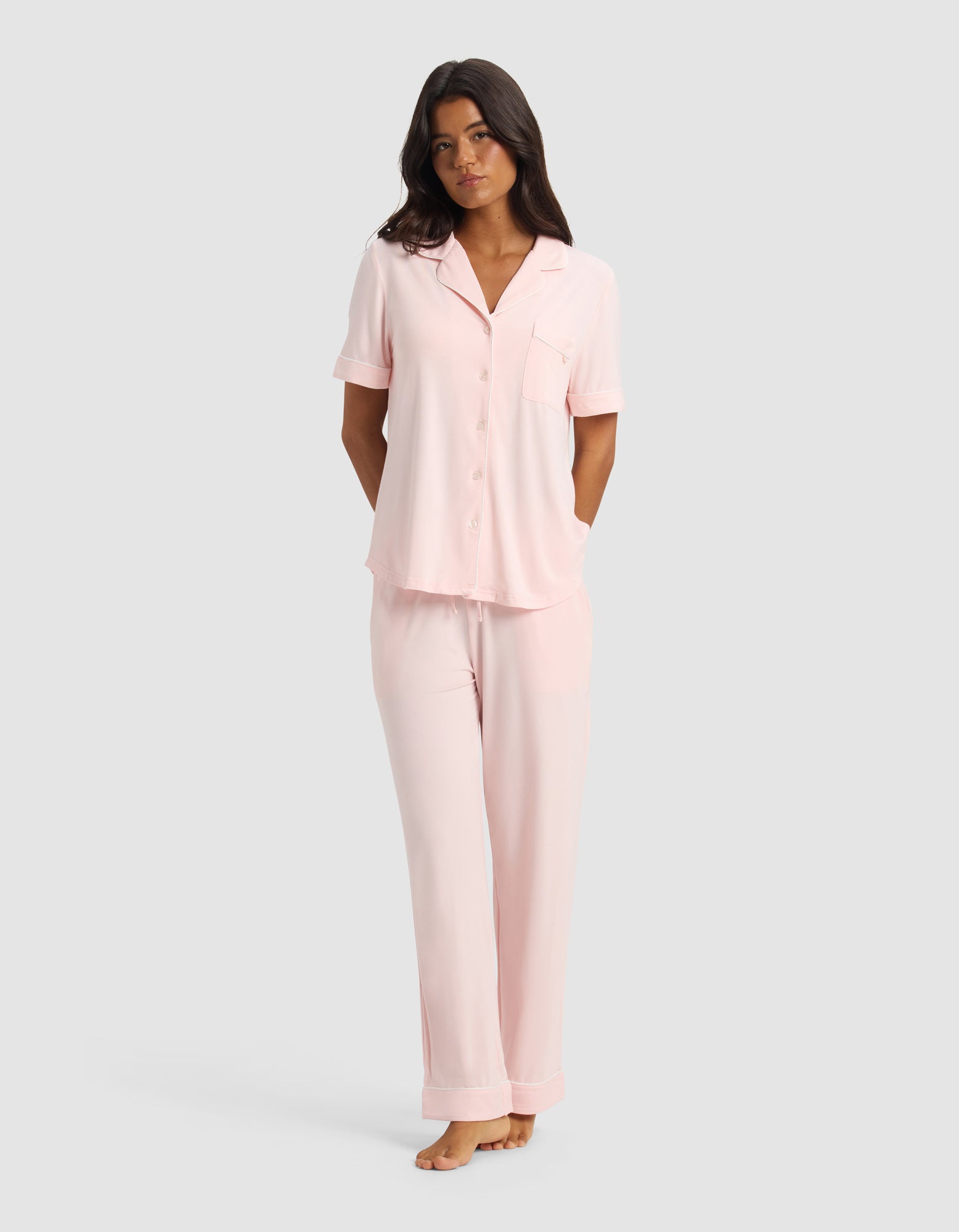 A woman stands barefoot against a plain light gray background, wearing the Cozy Earth Women's Bamboo Stretch-Knit Short Sleeve & Pant Pajama Set in Blossom, with her arms relaxed behind her back.