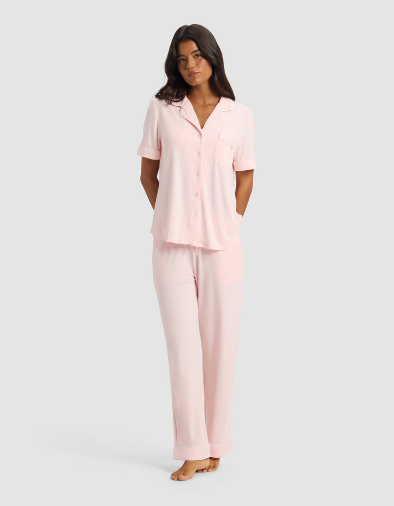 A woman stands barefoot against a plain light gray background, wearing the Cozy Earth Women's Bamboo Stretch-Knit Short Sleeve & Pant Pajama Set in Blossom, with her arms relaxed behind her back.
