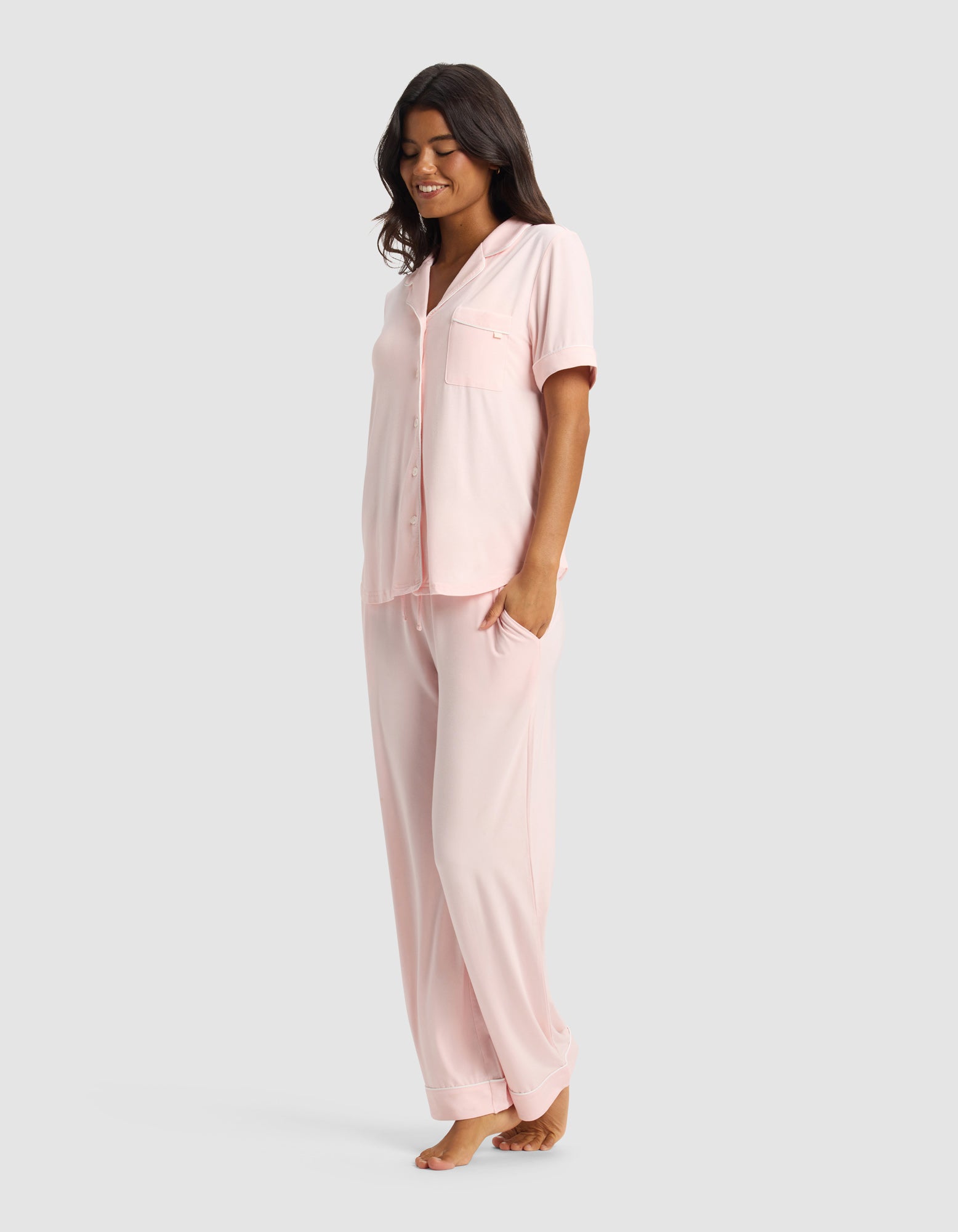 A woman stands barefoot, smiling, in the Cozy Earth Women's Bamboo Stretch-Knit Short Sleeve & Pant Pajama Set in light pink, with her left hand in her pocket against a plain light gray background.|Color:Blossom