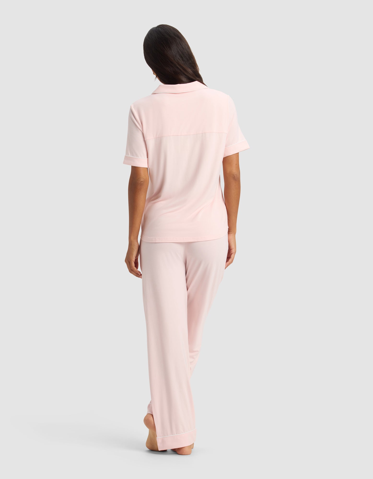 A woman stands barefoot, facing away, wearing the Cozy Earth Women's Bamboo Stretch-Knit Short Sleeve & Pant Pajama Set in light pink, against a plain gray background.