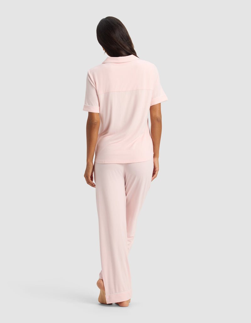 A woman stands barefoot, facing away, wearing the Cozy Earth Women's Bamboo Stretch-Knit Short Sleeve & Pant Pajama Set in light pink, against a plain gray background.