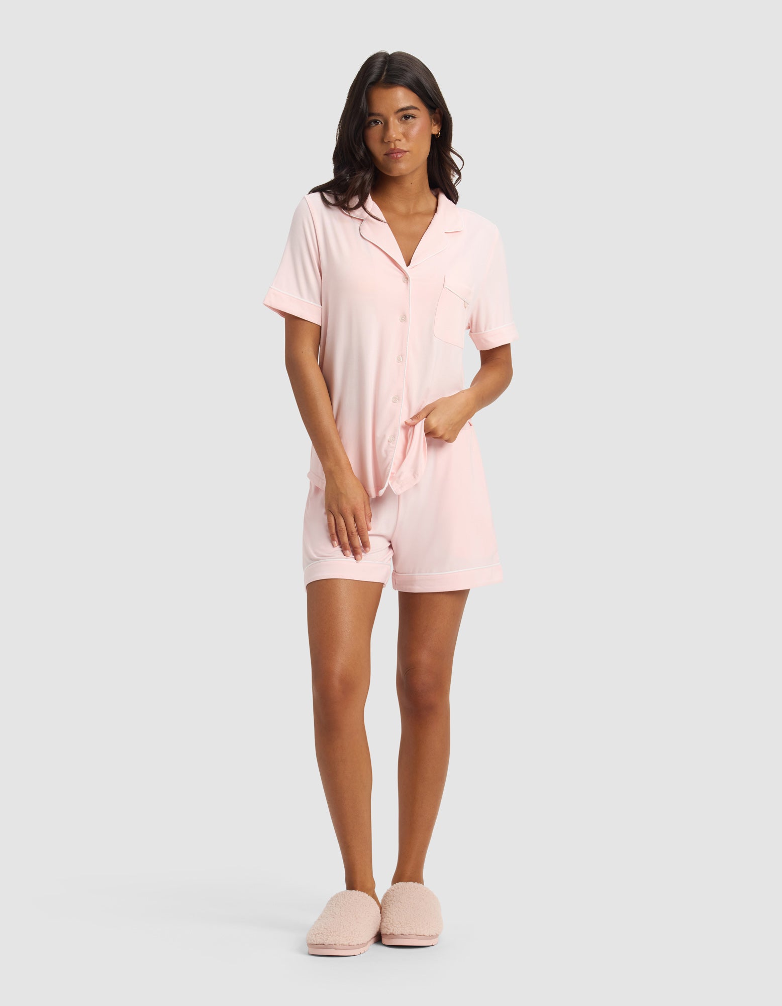 A woman faces forward wearing the Cozy Earth Women's Bamboo Stretch-Knit Short Sleeve Pajama Set in light pink with matching shorts and slippers, set against a plain light gray background.