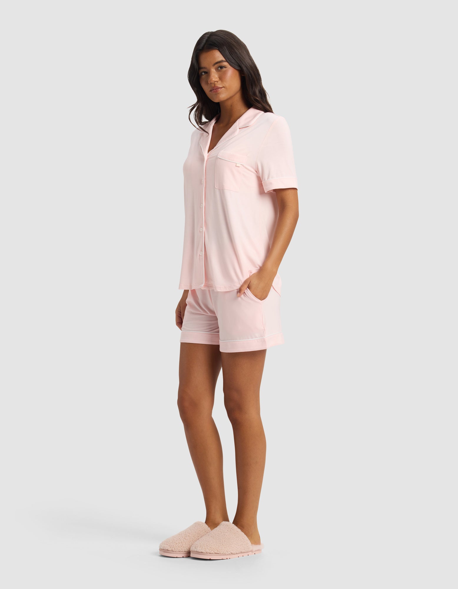 A woman wears the Cozy Earth Women's Bamboo Stretch-Knit Short Sleeve Pajama Set in pale pink with matching shorts and fluffy slippers, posing against a plain light gray background.|Color:Blossom