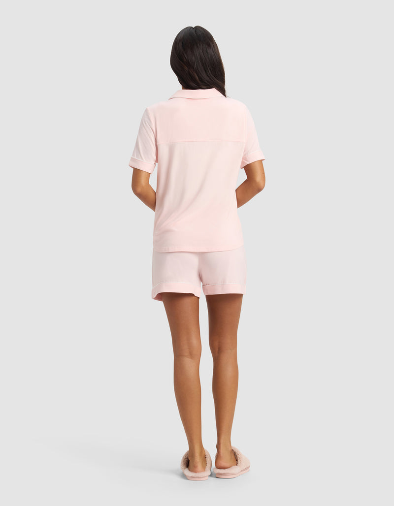 A woman faces away, wearing Cozy Earth’s Women's Bamboo Stretch-Knit Short Sleeve Pajama Set in light pink with matching shorts and pink slippers, set against a plain light gray background.